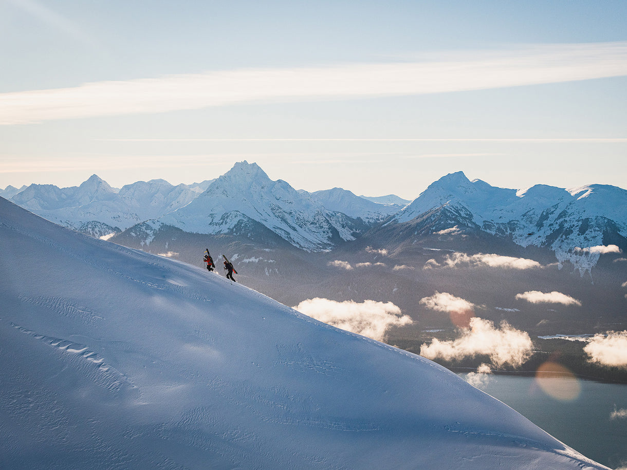 Ellen Bradley and Connor Ryan bootpack up a ridge near the ski area, with the peaks of Lingít Aaní (Tlingit land) towering in the distance.