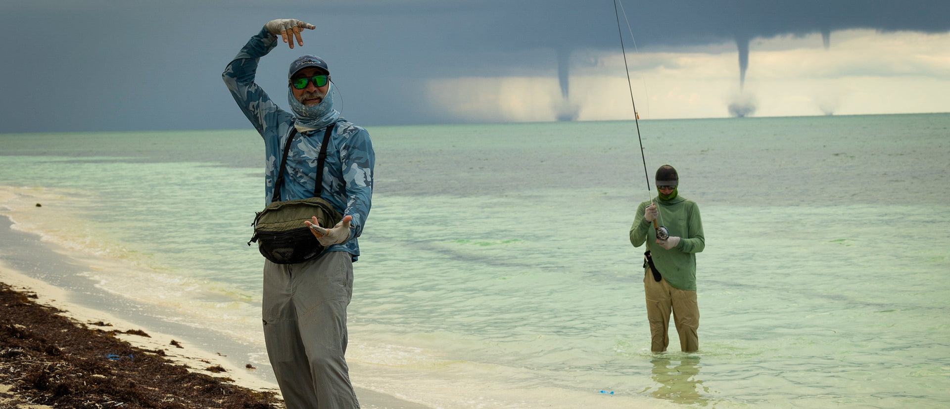 Two people fishing on a beach with clear water and a bridge in the background.