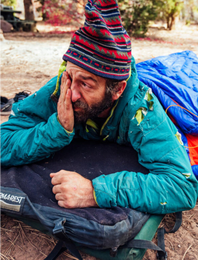 Man in teal jacket and colorful hat lying on a sleeping pad outdoors with a blue sleeping bag beside him.